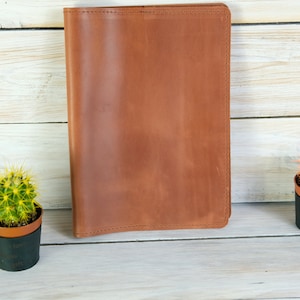 K&ouml;nnte beinhalten: Ein braunes Leder-Notizbuchcover mit einem gen&auml;hten Rand. Das Cover liegt auf einer wei&szlig;en Holzoberfl&auml;che mit zwei kleinen Topfkakteen auf jeder Seite.