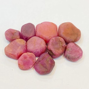 Op de afbeelding: Een verzameling gladde, gepolijste stenen in verschillende tinten roze en roodpaars. De stenen variëren in grootte en vorm, met een natuurlijk, getrommeld uiterlijk. Ze zijn gerangschikt tegen een witte achtergrond, waardoor hun kleuren en texturen worden benadrukt.