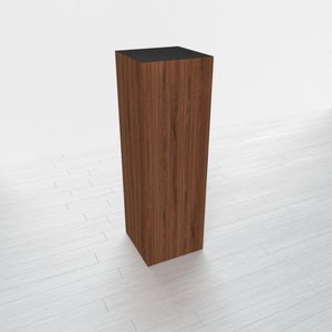 May include: A tall, rectangular wooden pedestal with a black top. The pedestal has a wood grain pattern and is standing on a white surface.