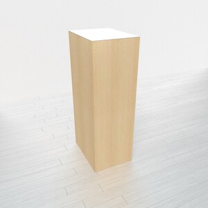 May include: A tall, rectangular wooden pedestal with a light wood finish. The pedestal is standing on a white wood floor.