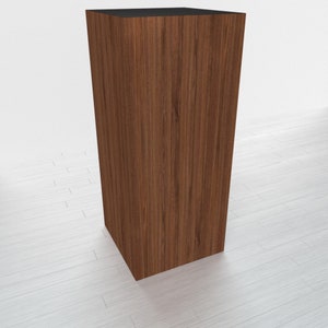 May include: A tall, rectangular wooden pedestal with a dark brown wood grain finish. The pedestal is standing on a white surface.