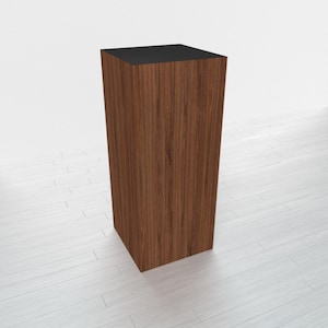 May include: A wooden pedestal with a black top. The pedestal is made of a dark wood with a grain pattern. It is a rectangular shape and is standing on a white surface.