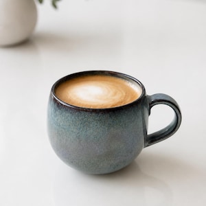 Op de afbeelding: Een ronde keramische mok met een gespikkeld blauw en grijs glazuur, gevuld met een koffiedrank met een witte schuimwerveling aan de bovenkant. De mok heeft een gebogen handvat en een afgeronde vorm met een verloop van donkere naar lichte tinten.