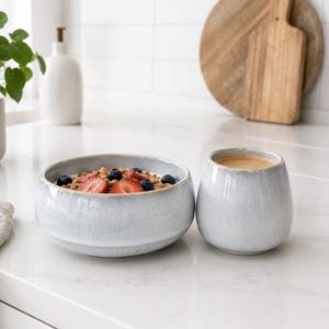 Puede incluir: Un cuenco de cerámica gris claro lleno de granola, fresas en rodajas y arándanos, junto a una taza de cerámica redondeada a juego que contiene una bebida de color marrón claro. Ambas piezas tienen un acabado liso y brillante.