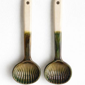 Può includere: Due cucchiai da portata in ceramica con manico color crema e coppetta verde scuro a forma di conchiglia. I manici hanno un piccolo foro in alto. Utensili da cucina decorativi.