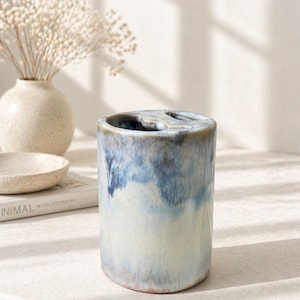 Op de afbeelding: Een cilindrische keramische tandenborstelhouder met een blauw-witte glazuur. De houder heeft een textuuroppervlak. Een kleine vaas met gedroogde bloemen en een kom staan op de achtergrond.