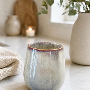 Op de afbeelding: Een keramieken beker met een afgerond, handvatloos silhouet. De buitenkant is voorzien van een reactief glazuur met een basis van zachte crème- en grijstinten, die overgaat in een opvallende, donkerpaars-bruine rand met een gladde afwerking.