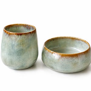 Op de afbeelding: Een keramische beker en een bijpassende ondiepe kom met een reactief glazuur in tinten saliegroen en blauw met een opvallende bruine rand. Beide stukken hebben een gladde, afgeronde vormgeving en een glanzende afwerking.