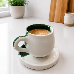 Op de afbeelding: Een ronde, witte keramische mok met een donkergroene binnenkant en handvat, gevuld met koffie. De mok staat op een bijpassende witte schotel met een donkergroen accent. Het ontwerp heeft een gladde, glanzende afwerking en een bolle vorm.