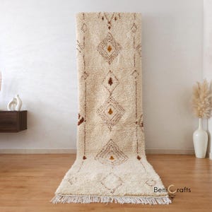 Op de afbeelding: Crèmekleurig, rechthoekig tapijt met een geometrisch ruitpatroon in bruin en goud. Het tapijt heeft een gefranjerde rand en wordt tegen een witte muur getoond. Het tapijt is gemaakt van een dik, getextureerd materiaal.