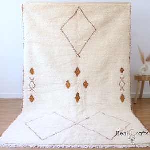 May include: A white Moroccan rug with a diamond pattern in a light brown color. The rug has a fringe border and the text "Beni Crafts" is visible in the bottom right corner.