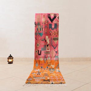 Könnte beinhalten: Ein langer, rechteckiger Teppich mit einem geometrischen Muster in Pink und Orange. Der Teppich hat an den Enden Fransen.