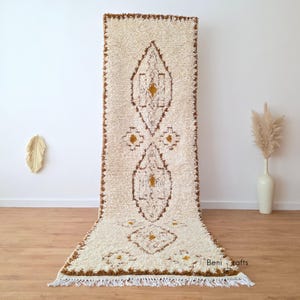Könnte beinhalten: Ein cremefarbener, rechteckiger Teppich mit einem braunen geometrischen Muster und Fransen. Der Teppich hat ein zentrales Rautenmuster mit kleineren Rauten- und Blumenakzenten. Die Kanten sind mit braunen Nähten versehen. Der Teppich wird auf einem Holzboden präsentiert.