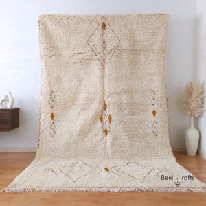 May include: Cream-colored, textured rug with a geometric diamond pattern in brown and gold. The rug has a fringe along the bottom edge and is displayed against a white wall. The rug is a floor covering.