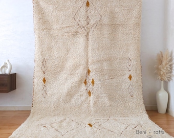 May include: Cream-colored, textured rug with a geometric diamond pattern in brown and gold. The rug has a fringe along the bottom edge and is displayed against a white wall. The rug is a floor covering.