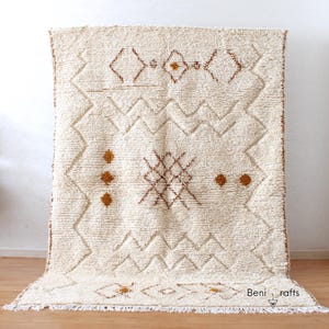 May include: Cream-colored, textured rug with geometric patterns in brown. The rug features diamond, zigzag, and dot designs. The rug has a fringed edge and is displayed on a wooden floor. The text "Beni rafts" is visible in the bottom right corner.