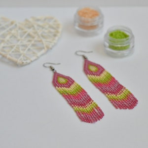 Könnte beinhalten: Ein Paar lange, rosa und grüne Perlenohrringe mit geometrischem Muster. Die Ohrringe hängen an silbernen Haken.