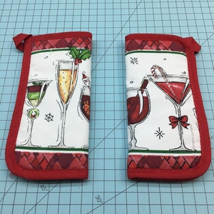 May include: Two red and white Christmas themed pot holders with a festive cocktail design. The pot holders feature a variety of cocktails, including a martini, a champagne flute, a margarita, and a glass of wine. The pot holders are perfect for adding a touch of holiday cheer to your kitchen.