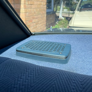 May include: A blue speaker grille with a circular pattern is mounted in the back of a car. The grille is made of metal and has a slightly textured surface.
