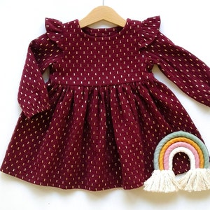 May include: A burgundy long-sleeved dress with gold dot accents, a ruffled neckline, and a gathered skirt. The dress is hanging on a wooden hanger. A colorful rainbow decoration with tassels is placed below the dress.