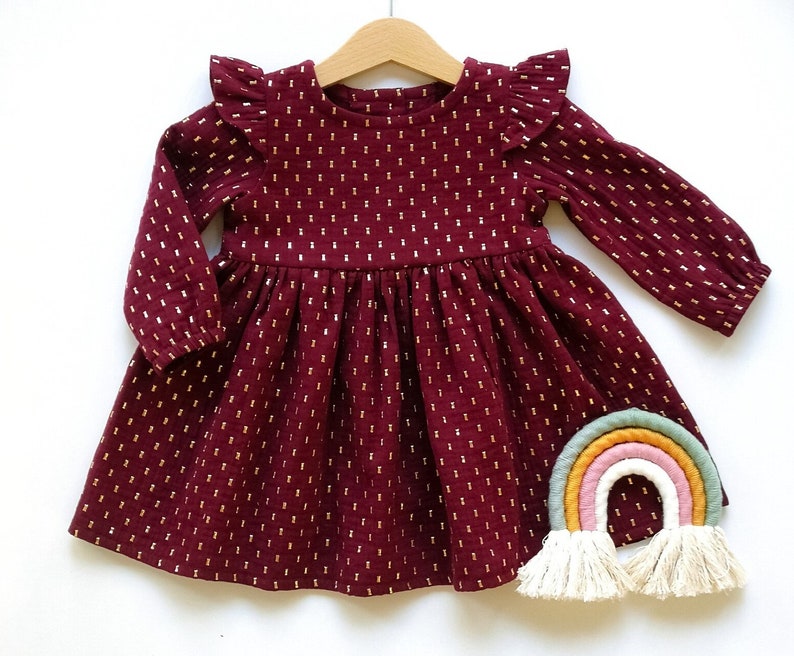 May include: A burgundy long-sleeved dress with gold dot accents, a ruffled neckline, and a gathered skirt. The dress is hanging on a wooden hanger. A colorful rainbow decoration with tassels is placed below the dress.
