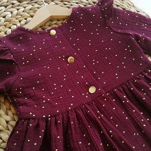 May include: A burgundy dress with gold polka dots, long sleeves, and a ruffled neckline. The dress features three gold buttons down the front and is hanging on a wooden hanger. The fabric appears to be a textured cotton.