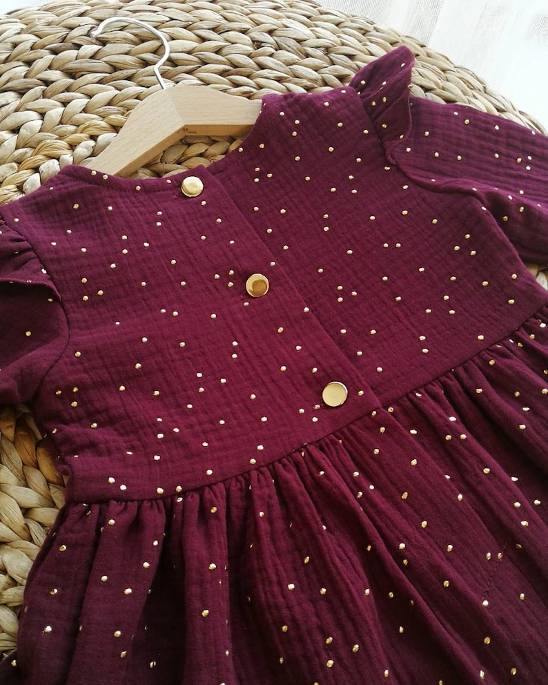 May include: A burgundy dress with gold polka dots, long sleeves, and a ruffled neckline. The dress features three gold buttons down the front and is hanging on a wooden hanger. The fabric appears to be a textured cotton.