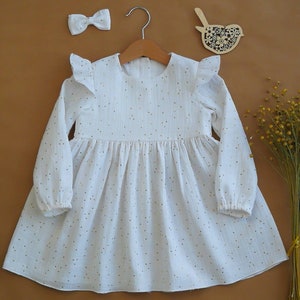 May include: A white long-sleeved dress with gold polka dots, a matching bow, and a wooden bird decoration. The dress has ruffled sleeves and is hanging on a wooden hanger.