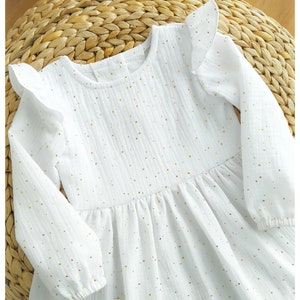 May include: White long-sleeved dress with gold polka dots and ruffle sleeves. The dress features a round neckline and a gathered waist. The fabric has a textured appearance. The dress is laying on a woven surface.