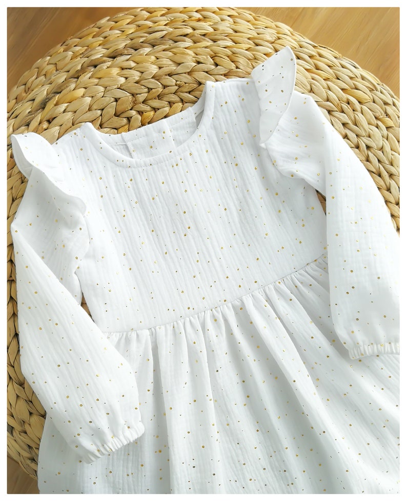 May include: White long-sleeved dress with gold polka dots and ruffle sleeves. The dress features a round neckline and a gathered waist. The fabric has a textured appearance. The dress is laying on a woven surface.