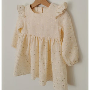 May include: A cream-colored baby dress with long sleeves and a gathered skirt. The dress features gold metallic polka dots and ruffled cap sleeves. The dress is hanging on a wooden hanger.
