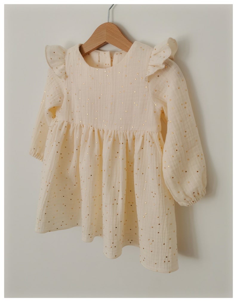 May include: A cream-colored baby dress with long sleeves and a gathered skirt. The dress features gold metallic polka dots and ruffled cap sleeves. The dress is hanging on a wooden hanger.