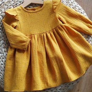 May include: Mustard yellow baby dress with long sleeves and a ruffled neckline. The dress is adorned with small, sparkling embellishments. The garment is hanging on a wooden hanger, resting on a woven surface.