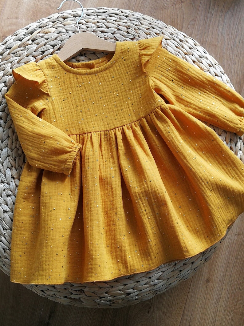 May include: Mustard yellow baby dress with long sleeves and a ruffled neckline. The dress is adorned with small, sparkling embellishments. The garment is hanging on a wooden hanger, resting on a woven surface.