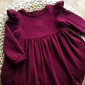 May include: A burgundy-colored long-sleeved dress with ruffled sleeves. The dress has a gathered waist and a round neckline. The fabric appears to be a textured, lightweight material. The dress is displayed on a woven, circular surface.