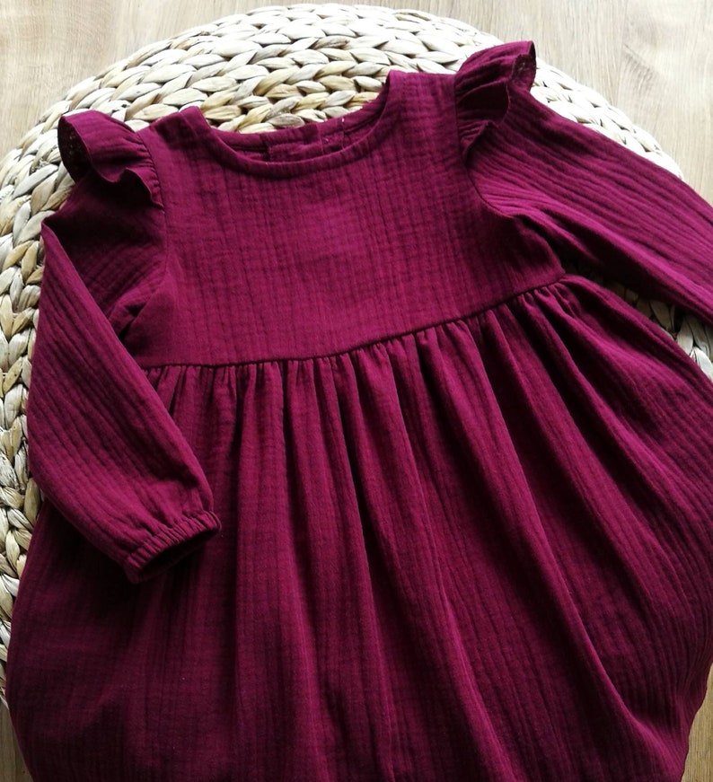 May include: A burgundy-colored long-sleeved dress with ruffled sleeves. The dress has a gathered waist and a round neckline. The fabric appears to be a textured, lightweight material. The dress is displayed on a woven, circular surface.