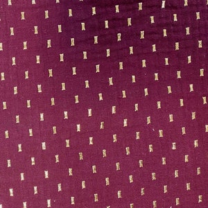 May include: Close-up of a burgundy fabric with a repeating pattern of small, rectangular gold accents. The gold shapes are evenly spaced across the surface, creating a textured appearance. The fabric has a subtle sheen.