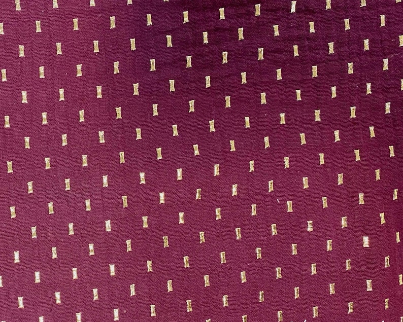 May include: Close-up of a burgundy fabric with a repeating pattern of small, rectangular gold accents. The gold shapes are evenly spaced across the surface, creating a textured appearance. The fabric has a subtle sheen.