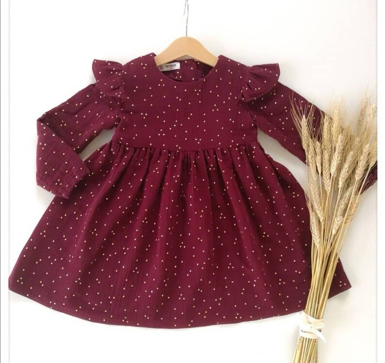 May include: A burgundy dress with long sleeves and a flared skirt, adorned with small white polka dots. The dress features ruffled sleeves and is hanging on a wooden hanger. A bundle of wheat stalks is placed to the right of the dress.
