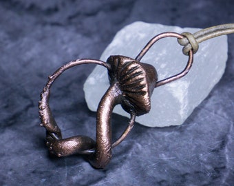 May include: A copper-coloured pendant featuring a mushroom design with a twisted stem. The pendant is hanging from a brown leather cord and is resting on a white crystal.