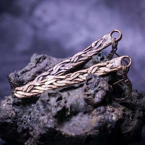 May include: A pair of copper earrings with a braided design. The earrings are on a dark gray textured surface.
