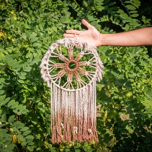 Op de afbeelding: Een bruine en witte droomvanger met een cirkelvormig geweven ontwerp en lange franjes die aan de onderkant hangen. De droomvanger wordt voor een groene, bladerrijke achtergrond gehouden.