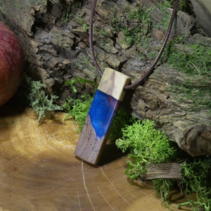 May include: A wooden pendant with a blue and purple resin inlay. The pendant is on a brown leather cord and is sitting on a wooden surface.