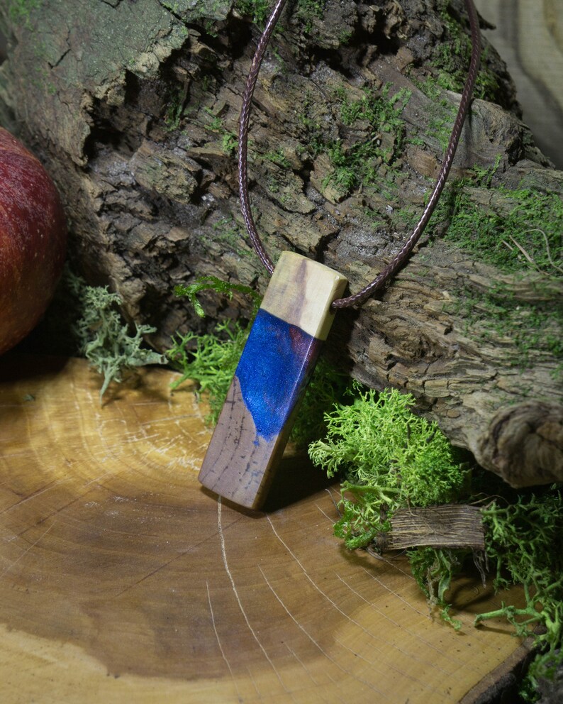 May include: A wooden pendant with a blue and purple resin inlay. The pendant is on a brown leather cord and is sitting on a wooden surface.