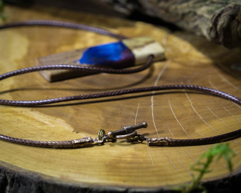 May include: A brown leather cord necklace with a wooden pendant featuring a blue and white resin inlay. The pendant has a natural wood grain pattern and a dark brown finish.