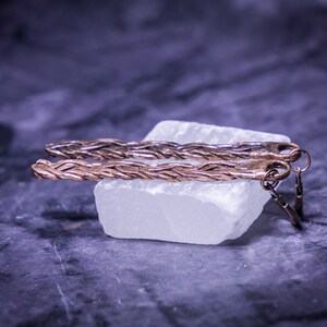 May include: A pair of long, braided copper earrings with a simple hook closure. The earrings are displayed on a white crystal.