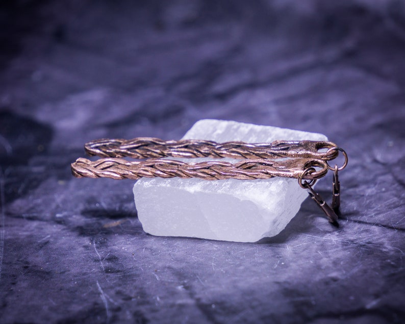 May include: A pair of long, braided copper earrings with a simple hook closure. The earrings are displayed on a white crystal.