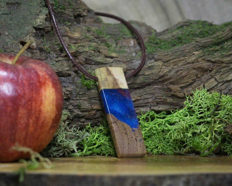 May include: A wooden pendant with blue and red resin inlay, hanging from a brown leather cord. The pendant is on a wooden surface with moss and a red apple.
