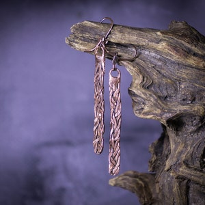 May include: A pair of long, copper earrings with a braided design. The earrings are hanging from a piece of driftwood.