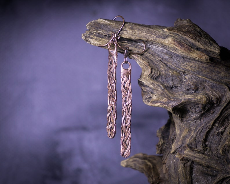 May include: A pair of long, copper earrings with a braided design. The earrings are hanging from a piece of driftwood.